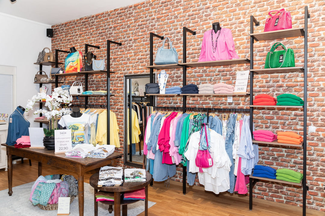 Modeauswahl bei Bella Vita in Miesbach – farbenfrohe Damenkleidung, edle Handtaschen und Accessoires vor rustikaler Backsteinwand.