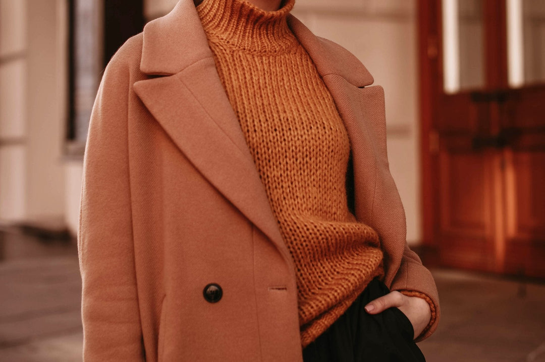 Frau in einem eleganten, camel-farbenen Wollmantel über einem grob gestrickten Rollkragenpullover in warmem Rostton – stilvoller Herbst-Look vor klassischem Stadthintergrund.