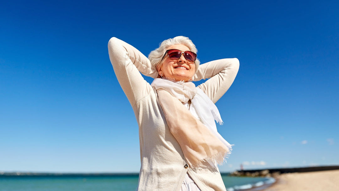 Ältere Frau genießt entspannt den Sonnenschein am Strand, trägt einen leichten, beigefarbenen Schal und Sonnenbrille – stilvoller Sommerlook mit Wohlfühlfaktor.