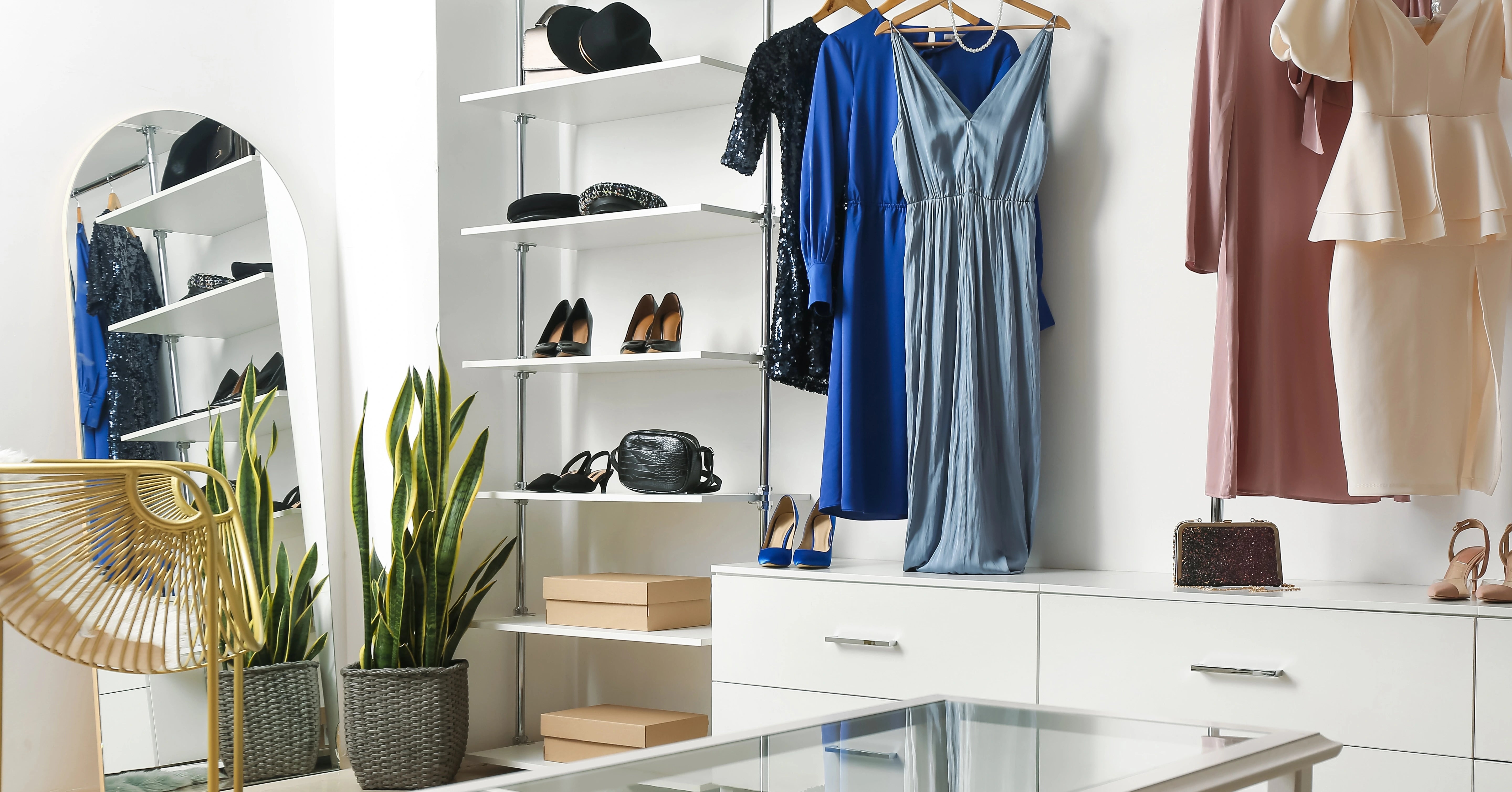 Heller Modeboutique-Innenraum mit Kleiderstange, eleganten Kleidern in Blau, Rosé und Beige, Schuhregal, Spiegel, Pflanzen und modernem Glastisch.