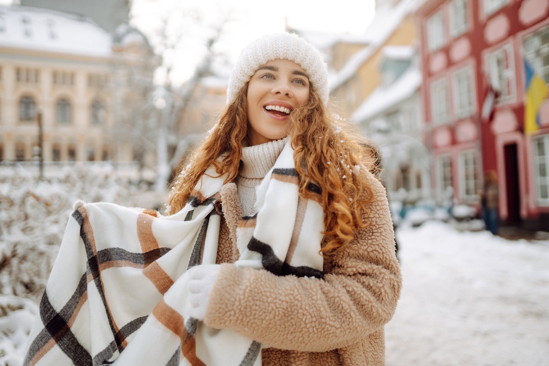 Fröhliche Frau mit kariertem Schal, Teddy-Mantel und Strickmütze im verschneiten Stadthintergrund – stilvolle Winteraccessoires bei Bella Vita – Mode & Lifestyle.